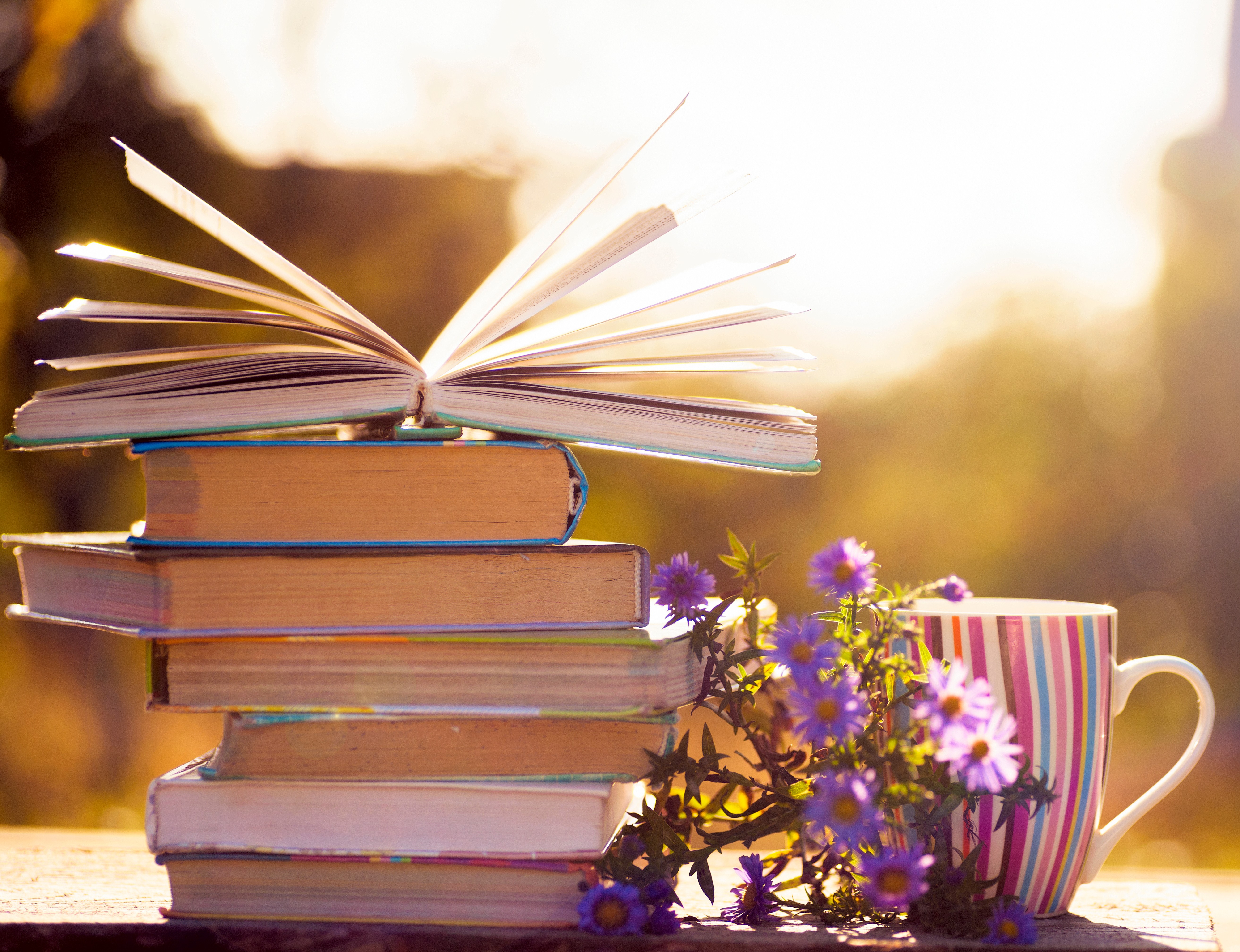 A stack of books next to a mug and a spring of purple flowers