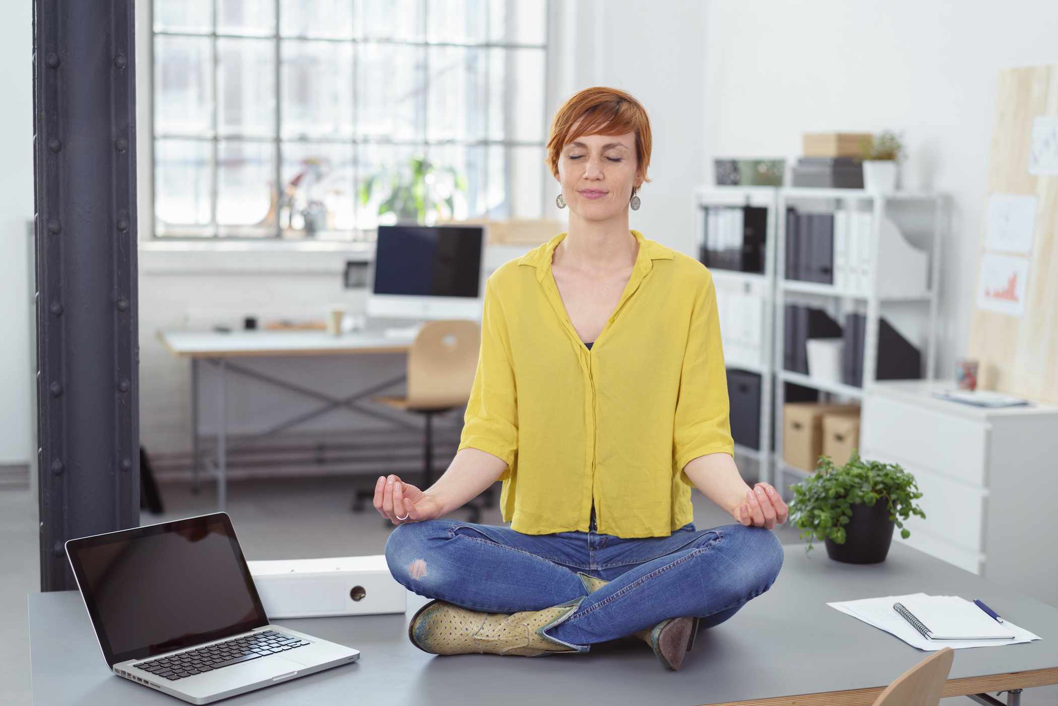A woman sits cross-legged in meditation on a desk in a bright office
