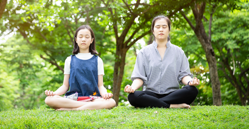 A parent and child sit side by side in meditation on the grass beneath trees