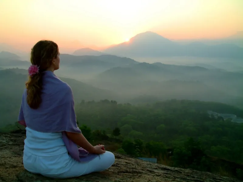 A woman sits in meditation on a hillside at sunrise, looking out over mist-filled valleys