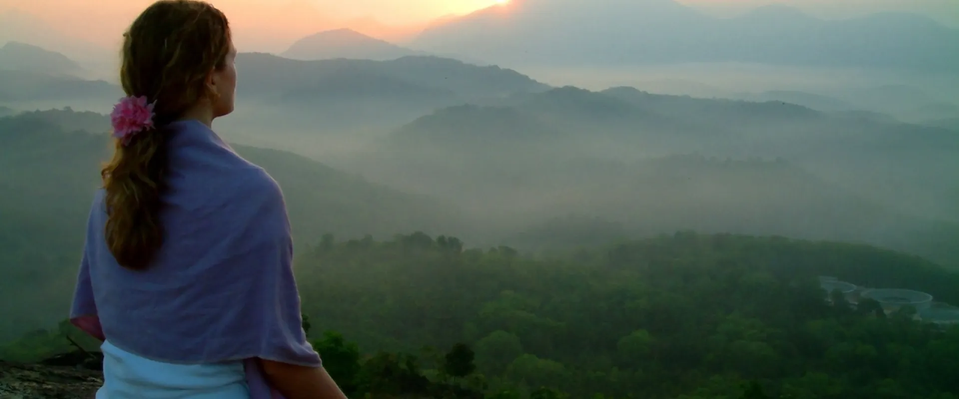 A woman sits in meditation on a hillside at sunrise, looking out over mist-filled valleys