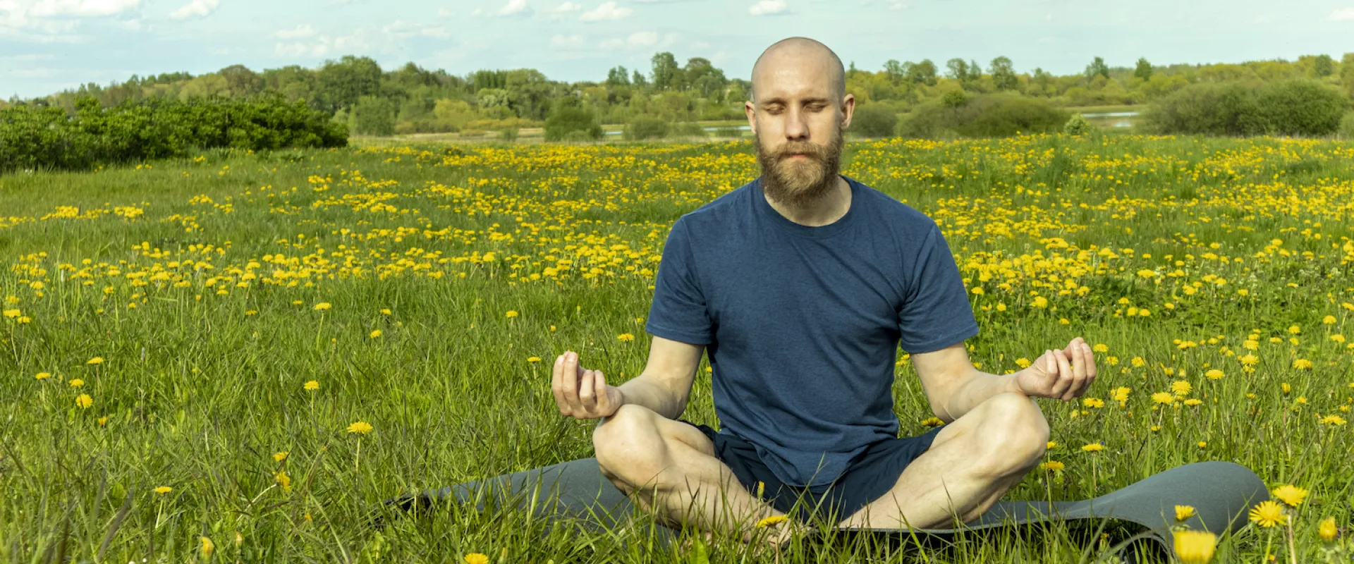 A man sits in meditation on a mat in a meadow of dandelions under a blue sky